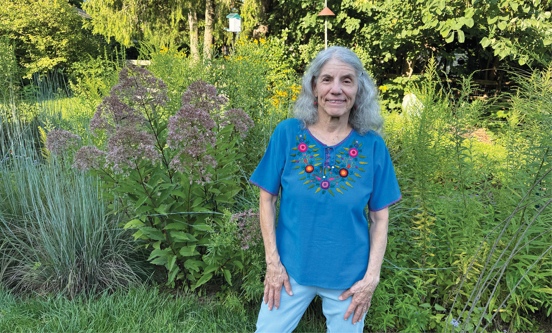 A photo of a women standing facing the camera in a blue shirt with jeans and gray hair.