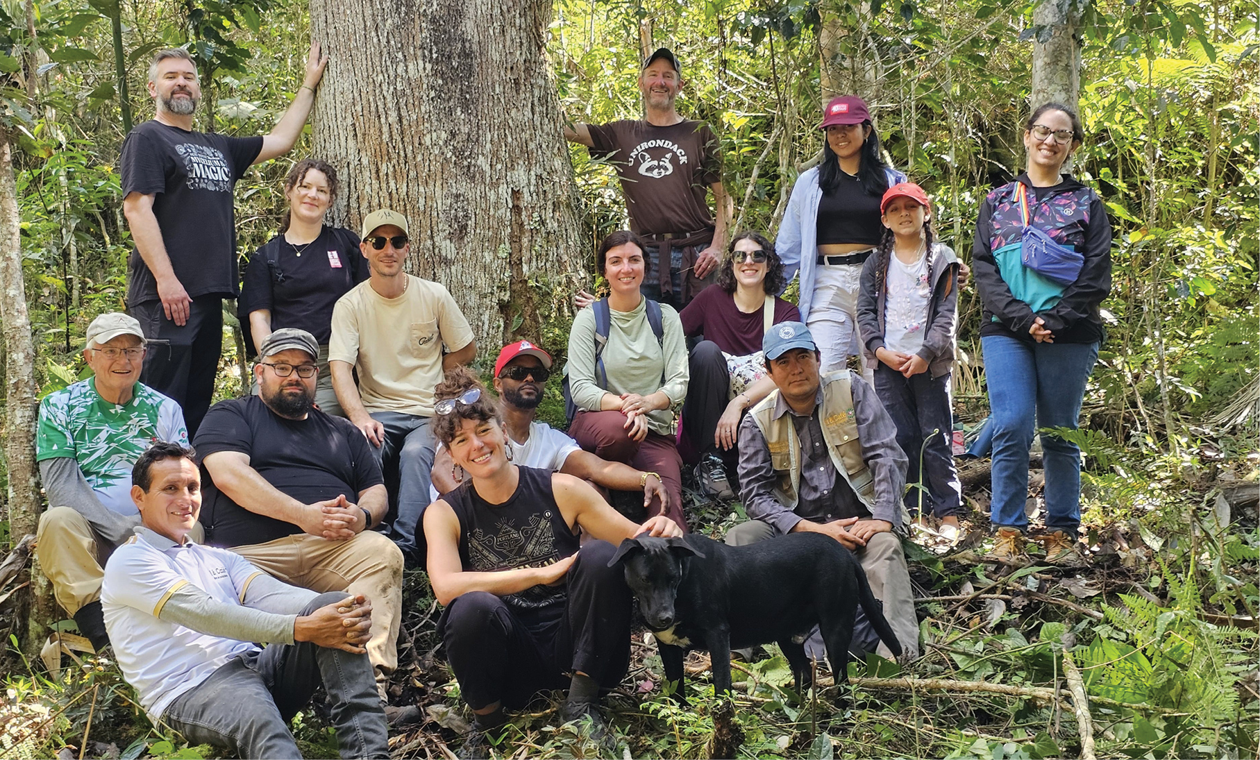 A group of people with a black dog sitting amongst the trees and smiling at the camera.